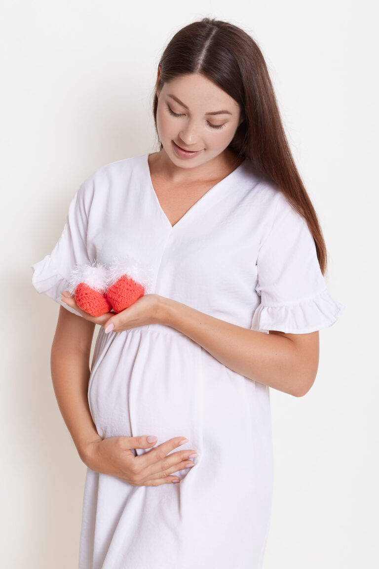 Gently pregnant woman with red booties, touching her belly, expectant mother with baby's booties posing against white wall, brunette lady with long beautiful expecting child Gently pregnant woman with red booties, touching her belly, expectant mother with baby's booties posing against white wall, brunette lady with long beautiful expecting child